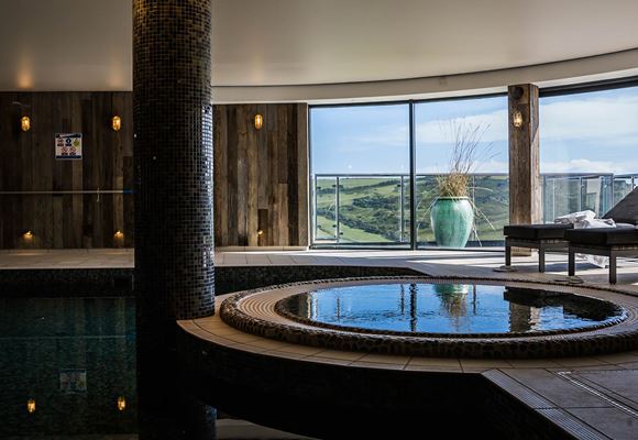 The indoor pool area at Gara Rock. Featuring wooden walls, a large central pillar, two loungers and views of the sea and surrounding greenery. 