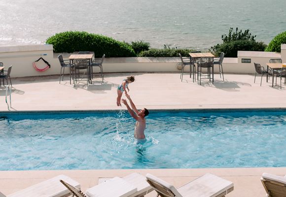 A father and daughter playing in the outdoor pool at Gara Rock with glorious sea views in the background.